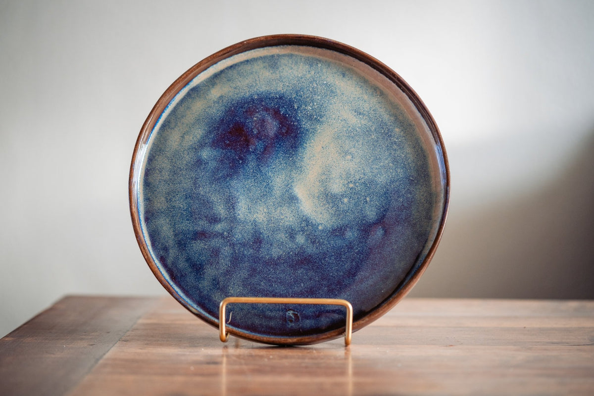 Blauw geglazuurd keramieken dinerbord met paarse vlekken op een standaard.