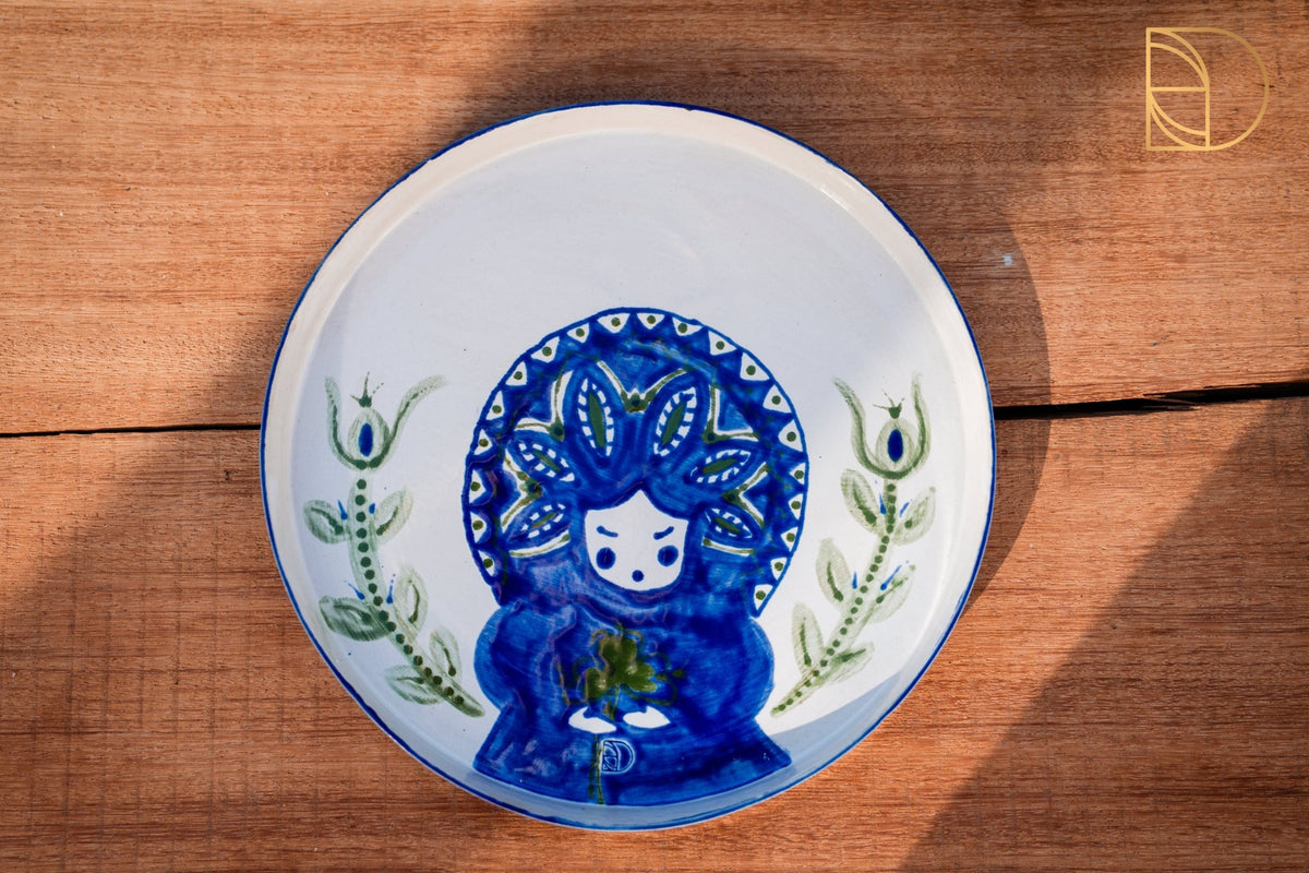 Wit keramieken bord met een blauw geschilderd folkloristisch figuur en groene bladranken.