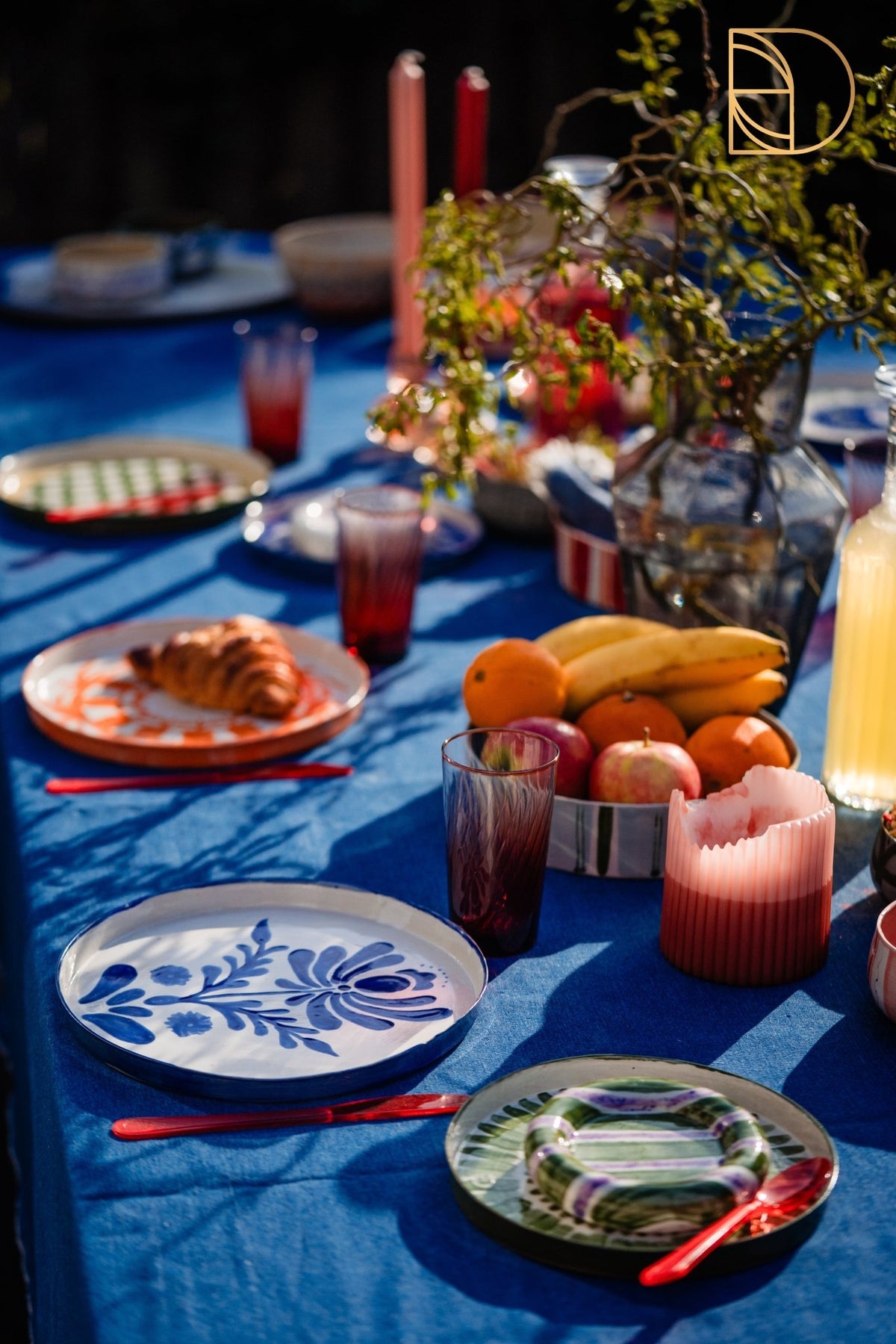 Gedekte tafel met blauw kleed, fruit, croissants en handbeschilderde borden.