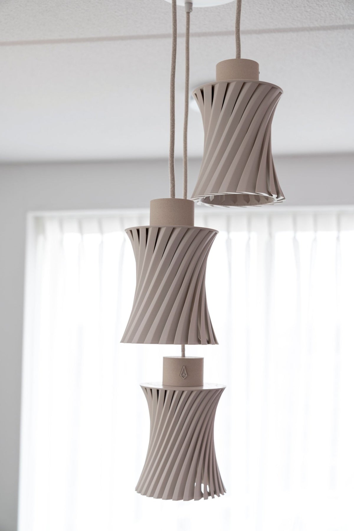 Hanglamp met drie zandbeige geribbelde kappen onder elkaar aan katoenen snoeren voor een raam met witte gordijnen.