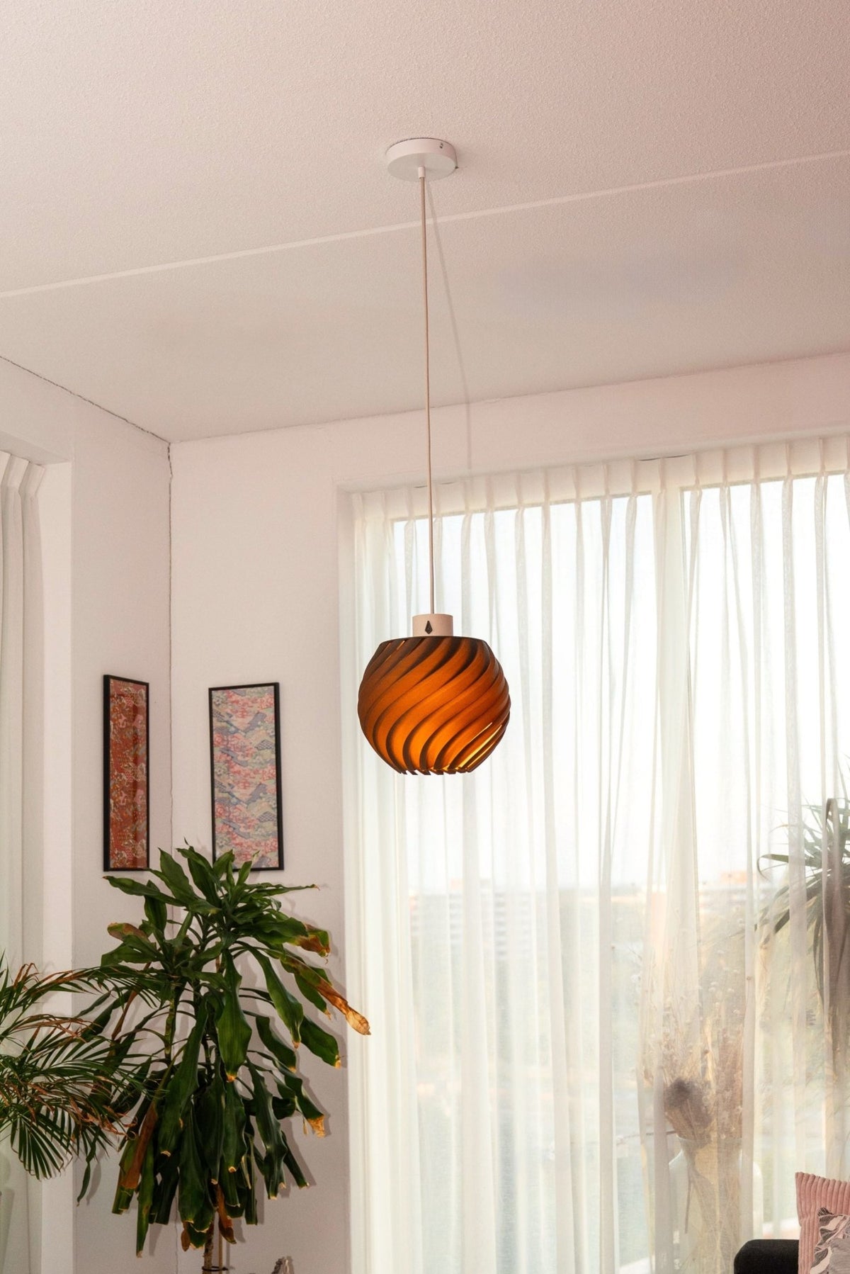 Brandende bolvormige hanglamp in een lichte kamer met een grote plant en twee lijsten aan de wand.