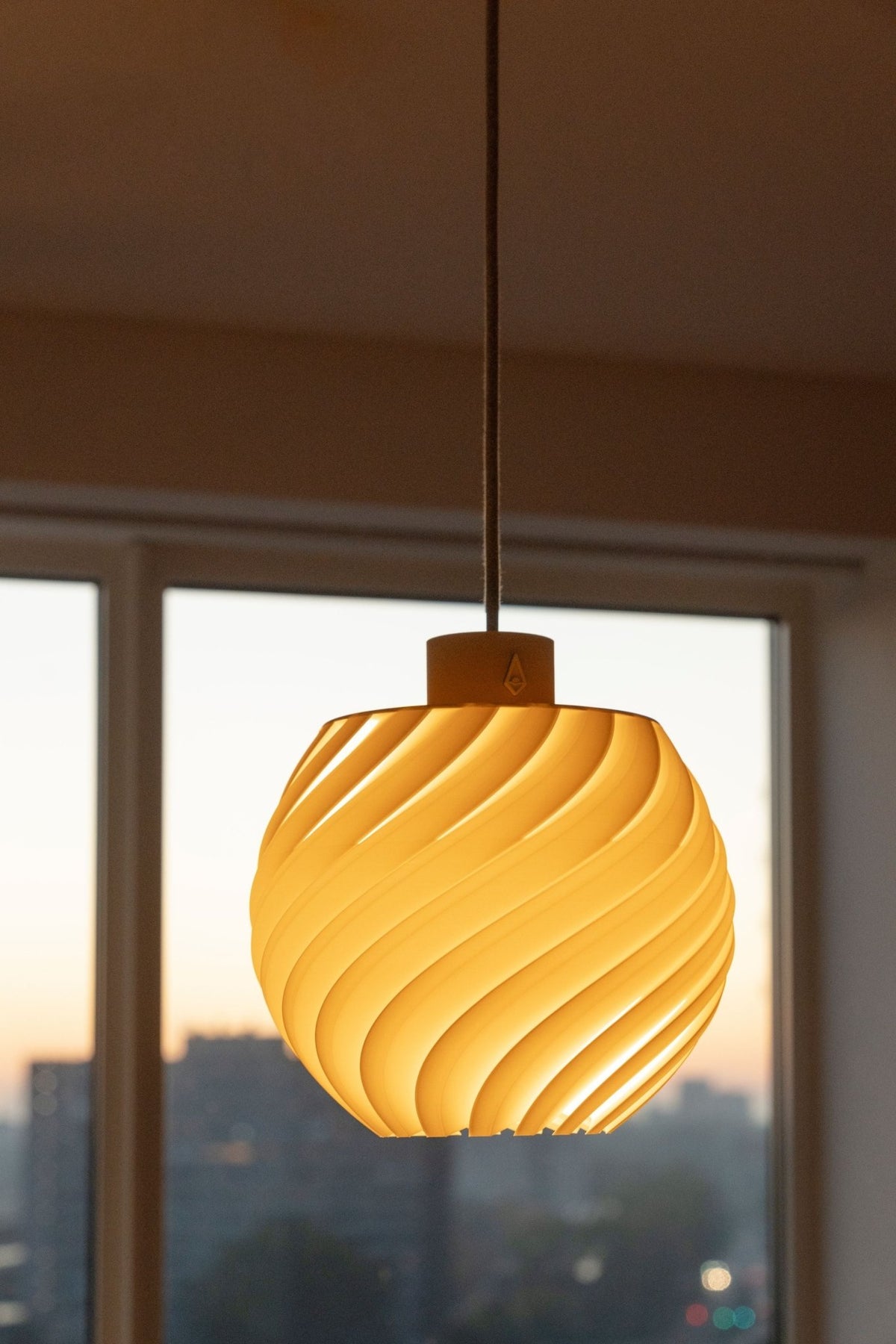 Warm oplichtende bolvormige hanglamp voor een raam met uitzicht op de stad bij zonsondergang.