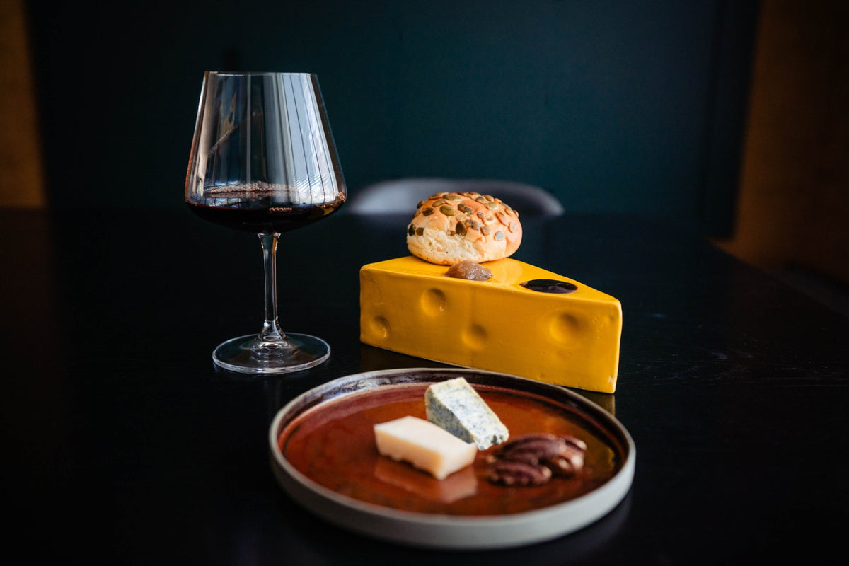 Gele keramieken kaaswig met een broodje erop, naast een glas rode wijn en een schaaltje kaas.