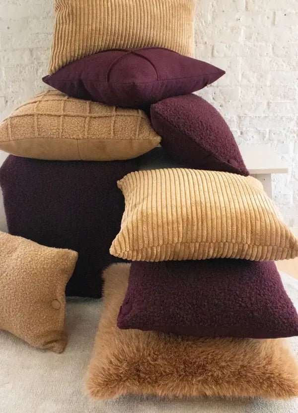 Stapel kussens in aubergine en camel van bont, bouclé en ribstof tegen een witte muur.