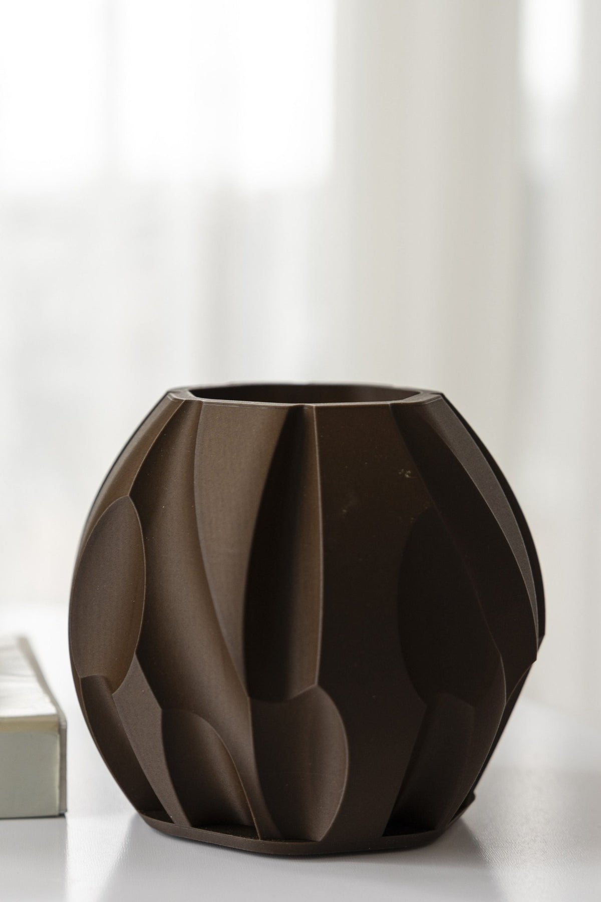Donkerbruine compacte tafellamp met facetreliëf op een witte tafel voor een licht gordijn.