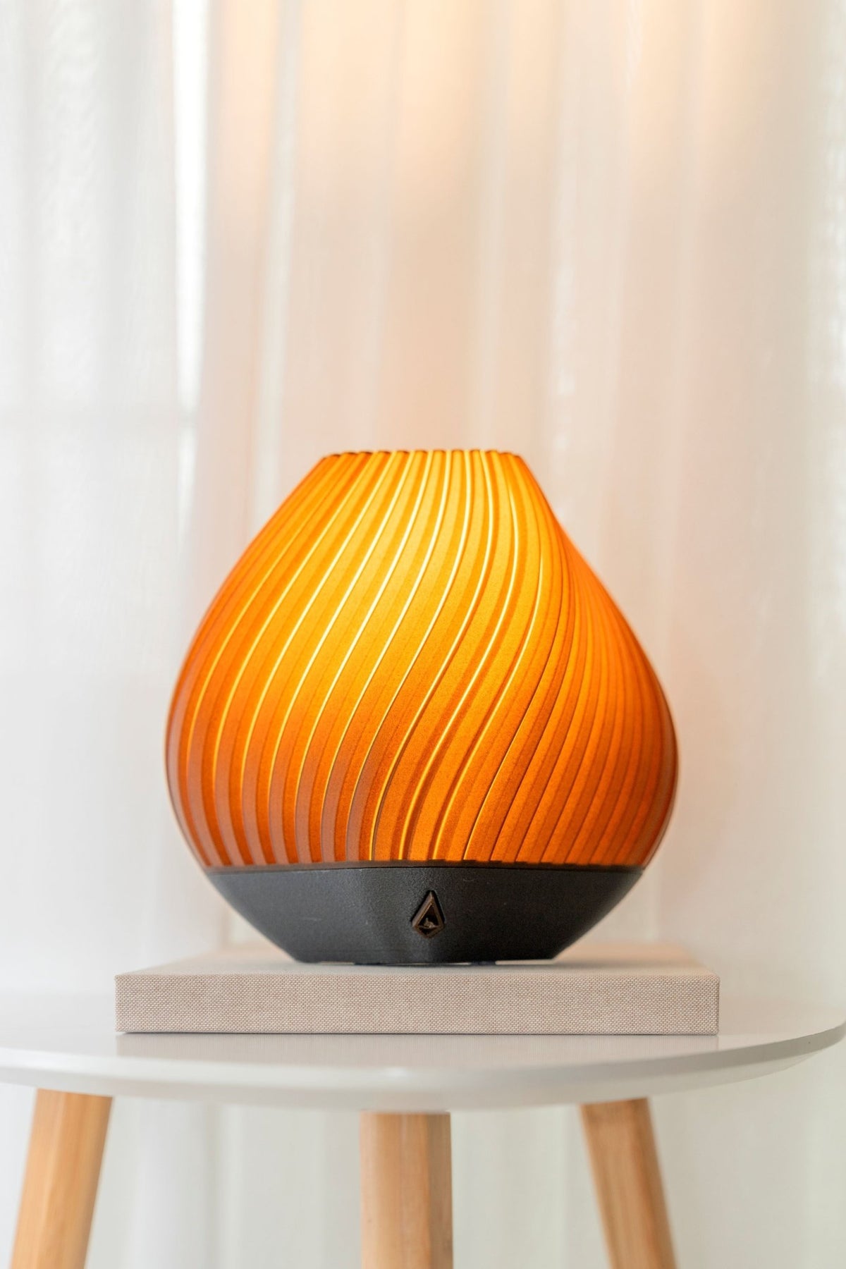 Geribbelde ronde tafellamp met brandend licht op een houten voet, op een bijzettafel voor een wit gordijn.