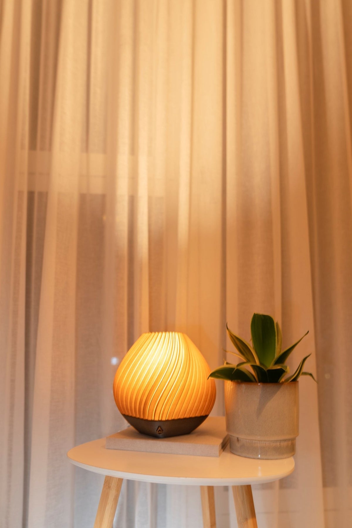 Brandende ronde tafellamp naast een plantje op een witte bijzettafel voor een warm verlicht gordijn.