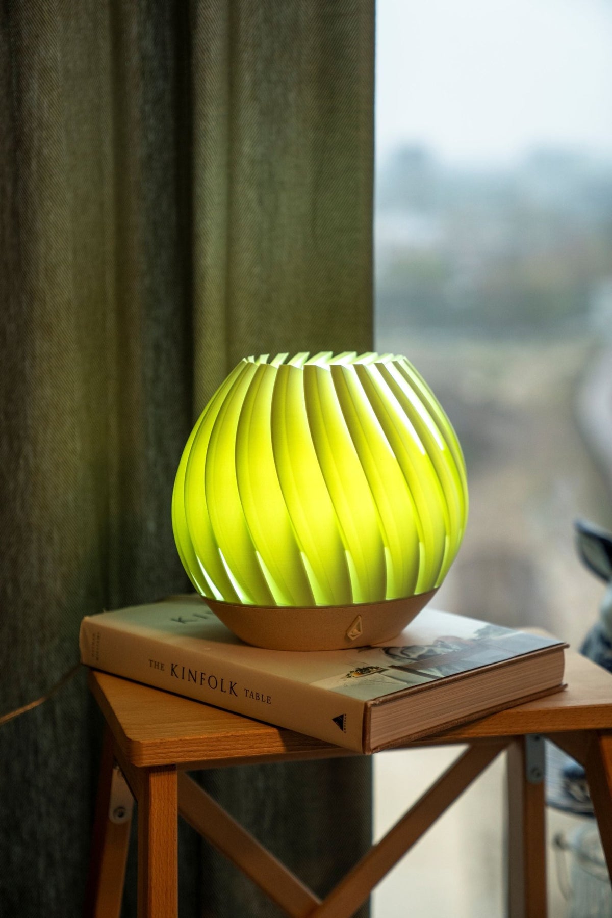 Fel groen oplichtende brede tafellamp op een boek op een houten bijzettafel bij een raam.