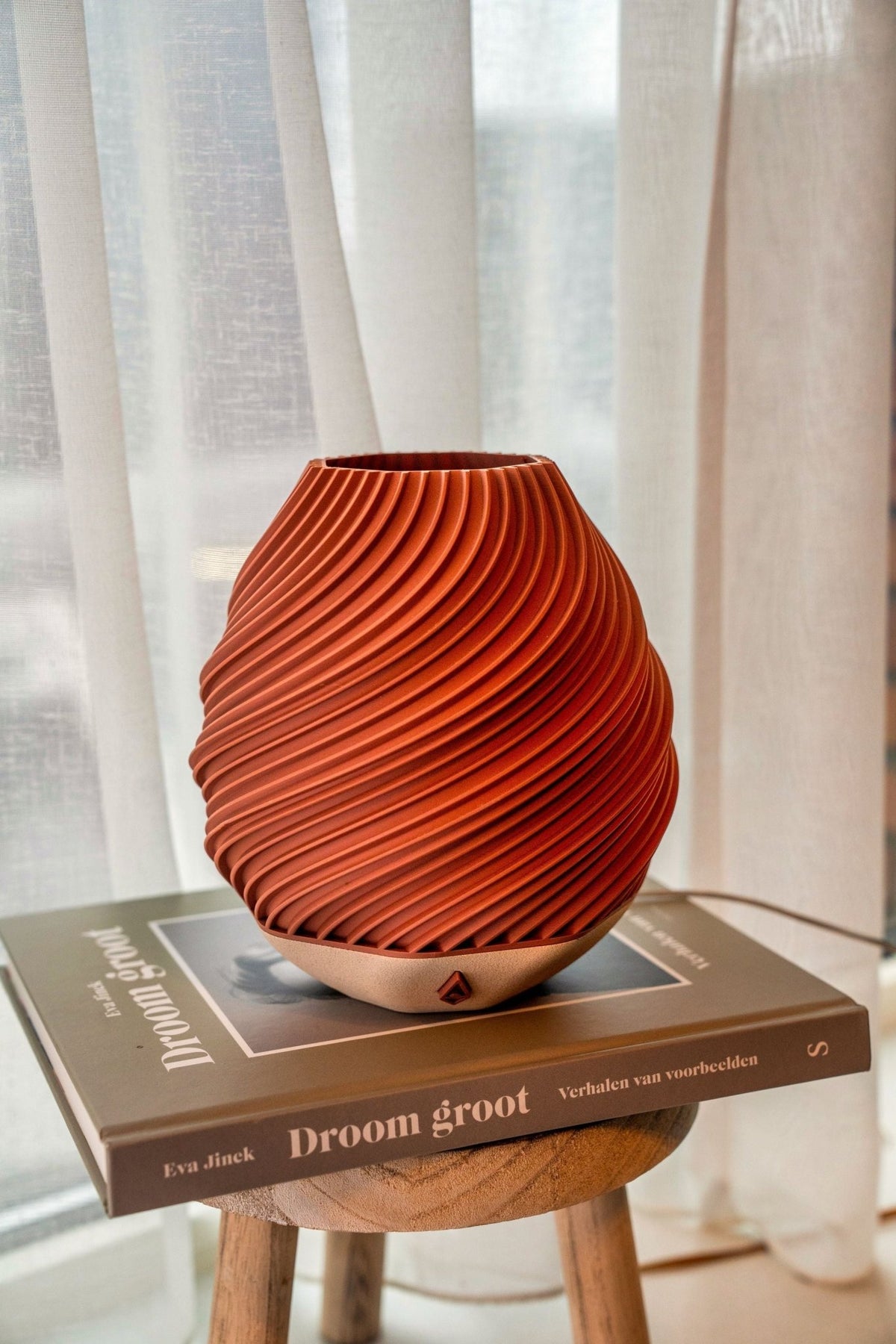 Baksteenrode gedraaide tafellamp op een boek op een houten krukje voor een licht gordijn.