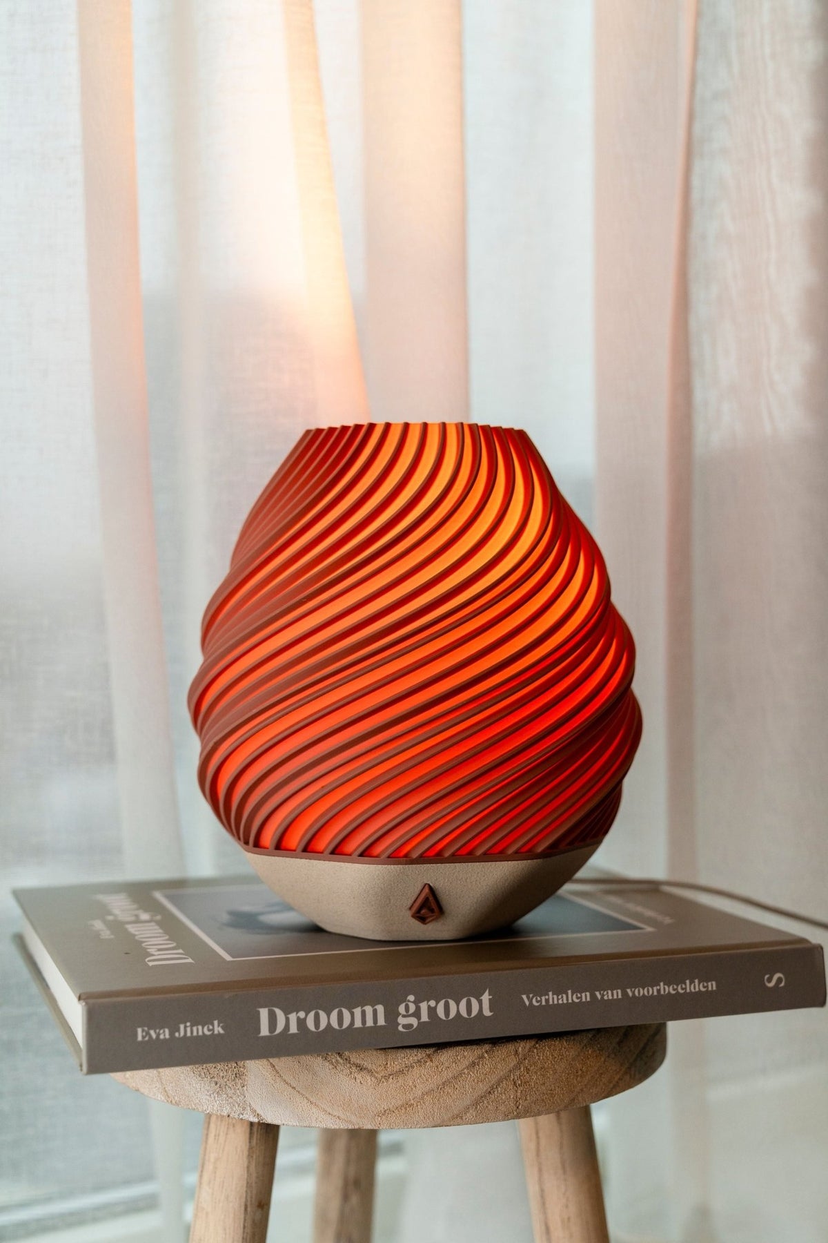 Baksteenrode tafellamp met gedraaide ribbels op een houten krukje voor een licht gordijn.