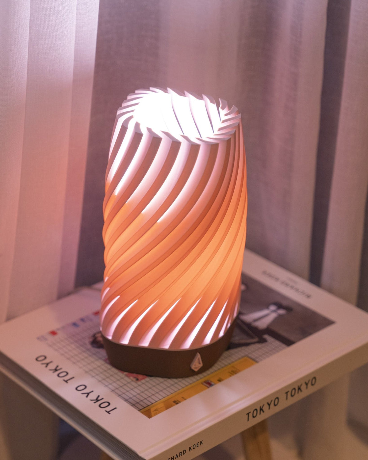 Koraalroze smalle gedraaide tafellamp met brandend licht op een boek voor een roze gordijn.