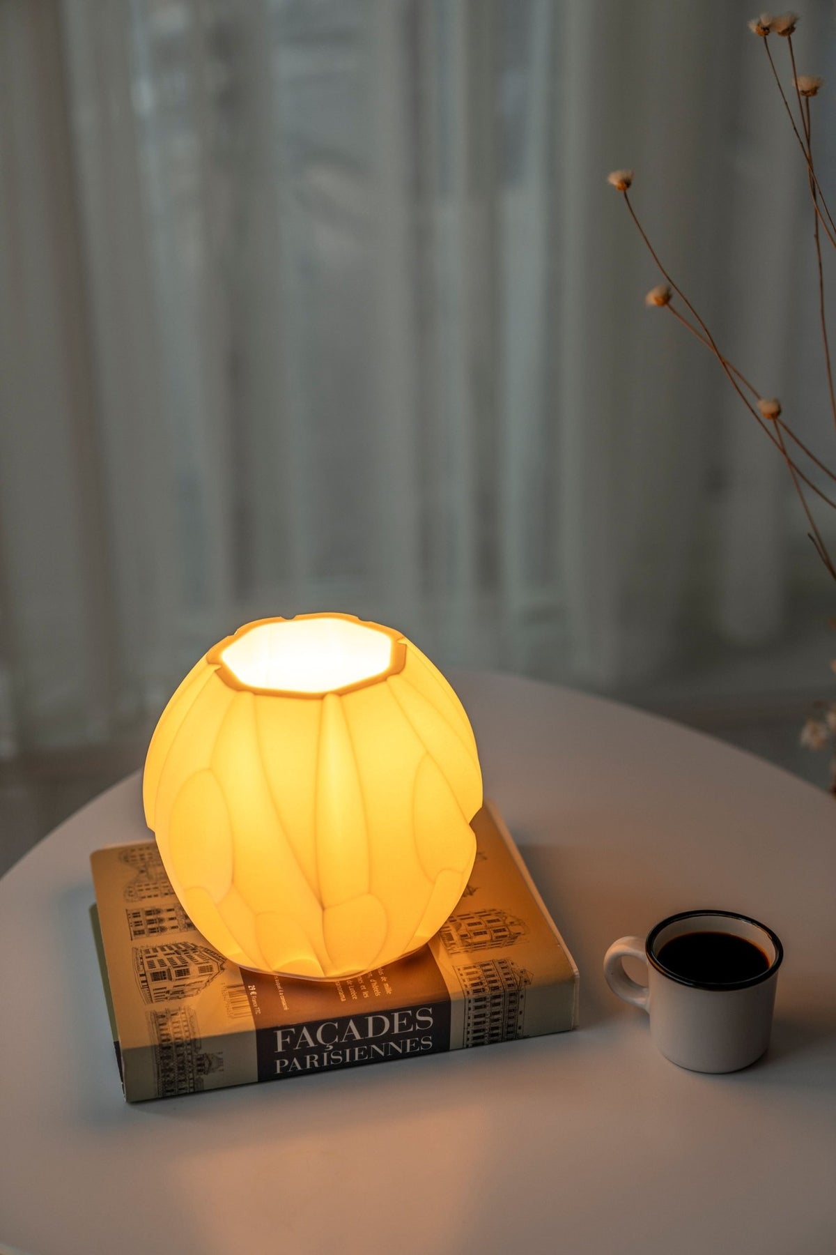 Gebroken witte compacte tafellamp met warm brandend licht op boeken naast droogbloemen.