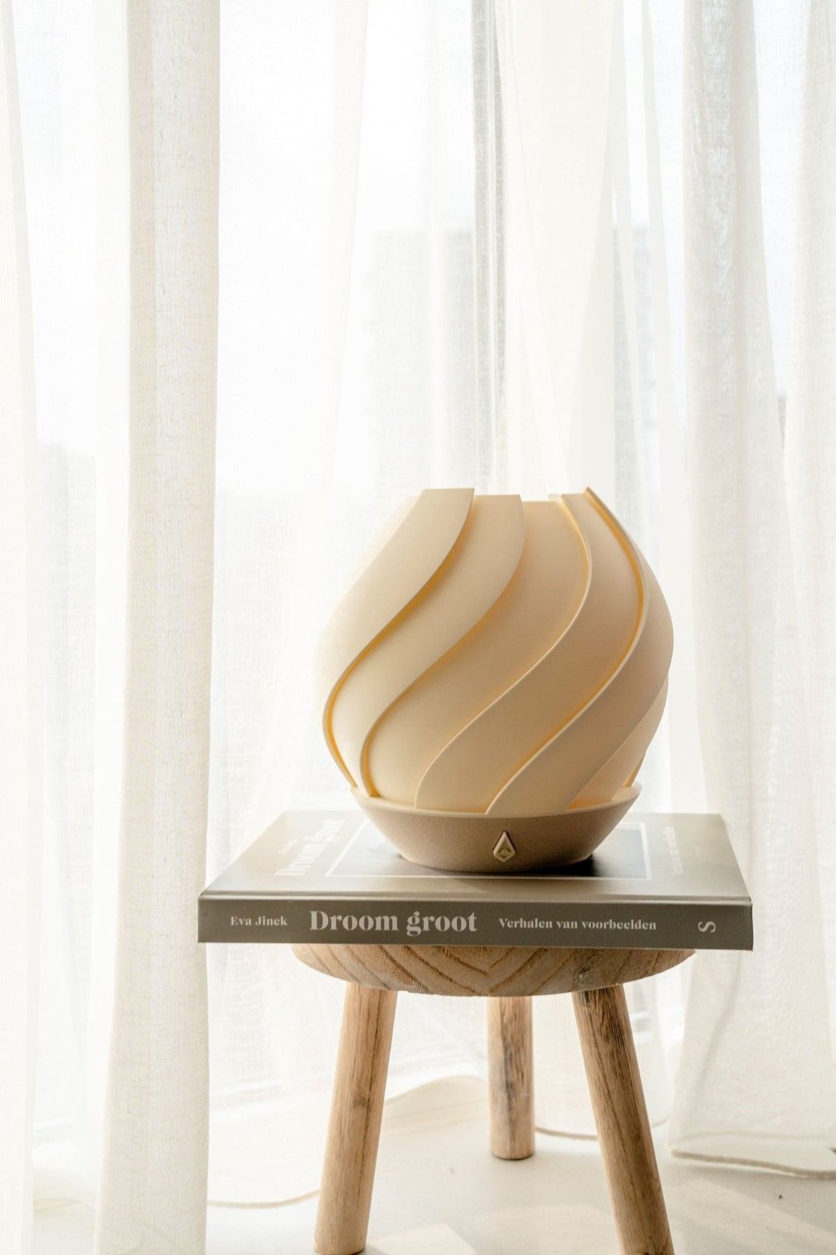 Gebroken witte bolvormige tafellamp met zeewierreliëf op een houten krukje voor een wit gordijn.