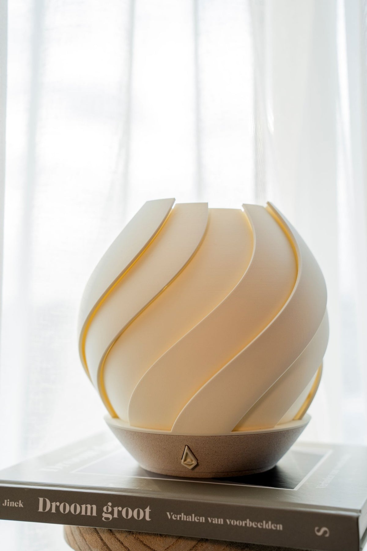 Gebroken witte bolvormige tafellamp met overlappend zeewierreliëf op een houten voet, op een boek.