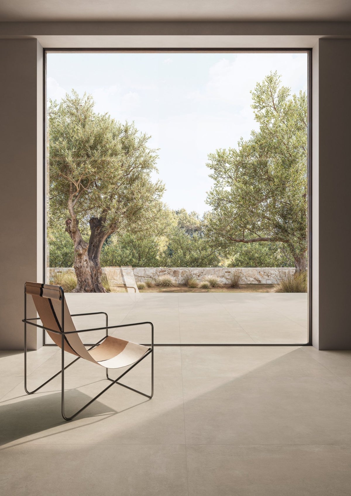 Grijsbeige betonlook vloer bij een openslaande pui, met leren fauteuil en uitzicht op olijfbomen.
