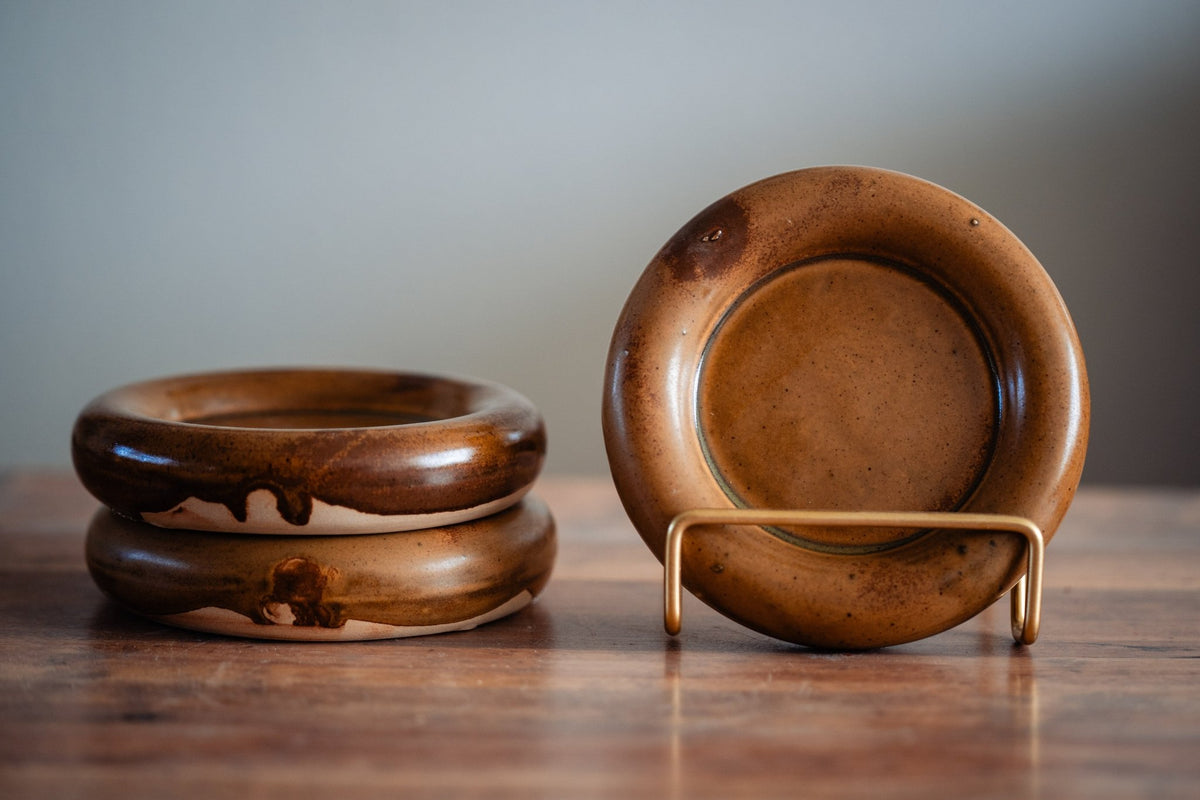 Klein rond keramieken bordje met dikke opgerolde rand in karamelbruin glazuur, met een tweede op een standaard.