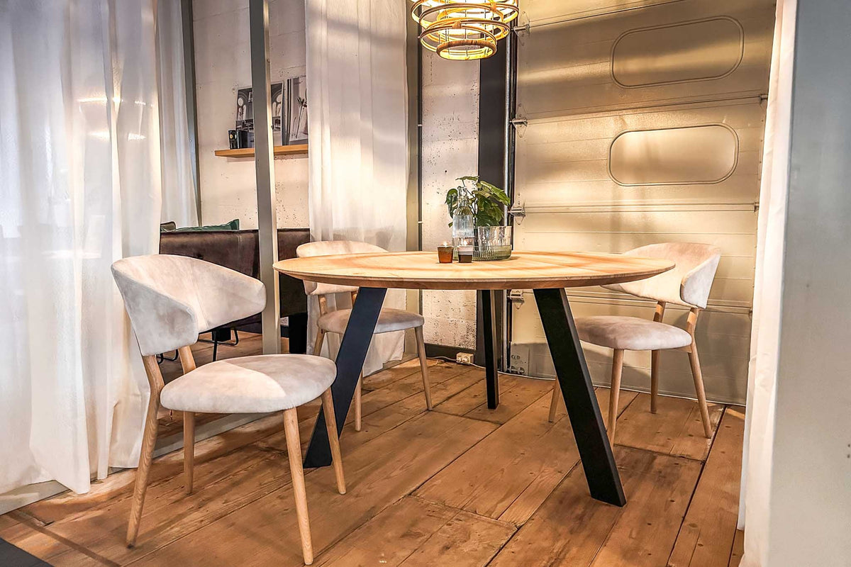 Ronde eiken eettafel op een zwarte stalen driepoot, met lichte stoelen op een oude houten vloer