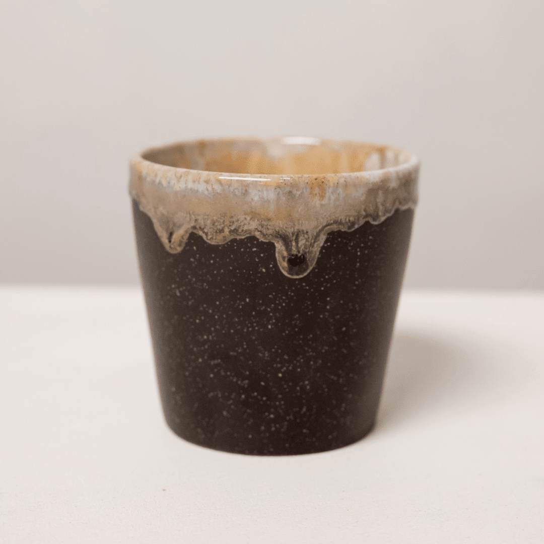 Klein donkerbruin keramieken kopje zonder oor met crème druipglazuur langs de rand.