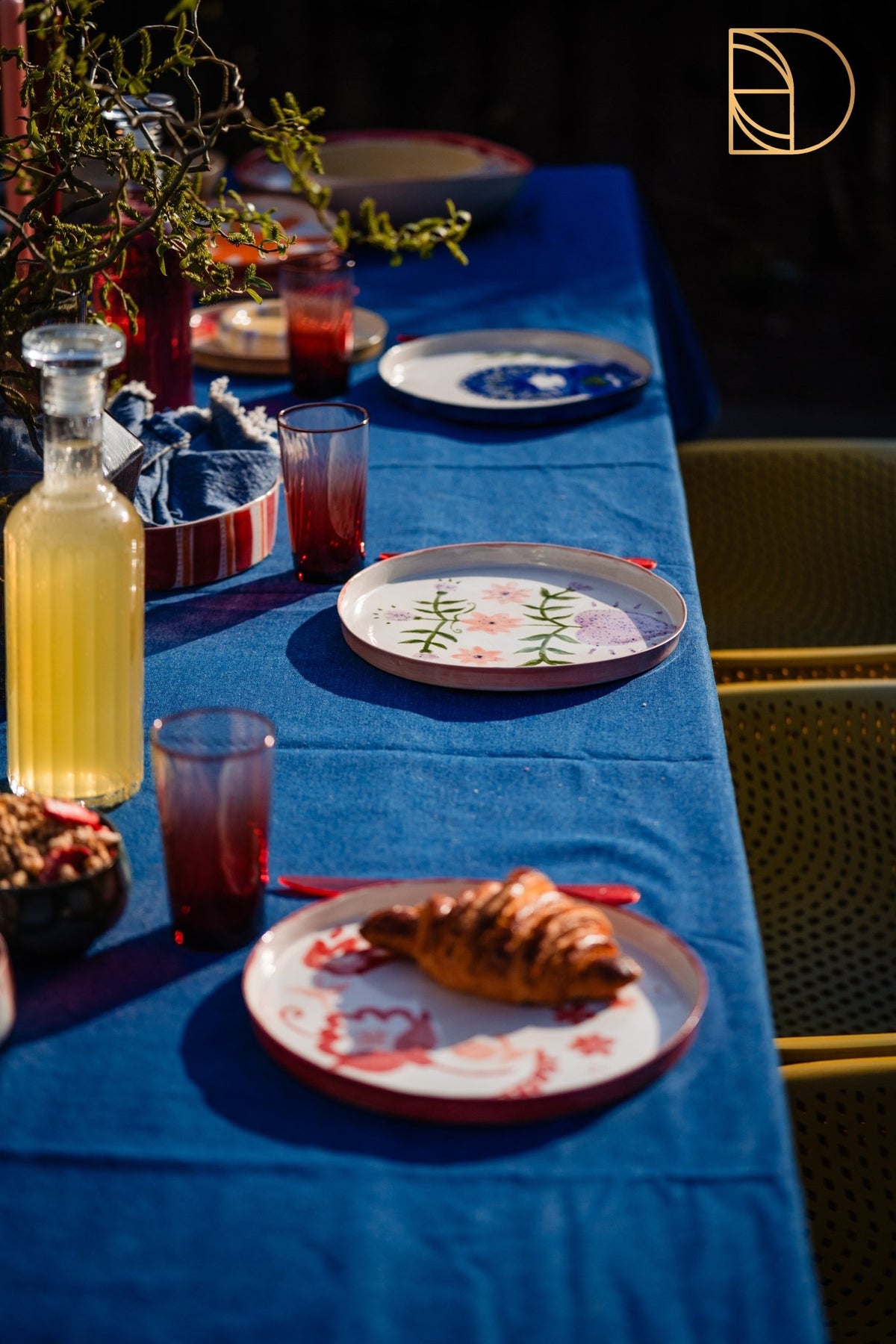 Lange gedekte tafel met blauw kleed en handbeschilderde borden met bloemmotieven.