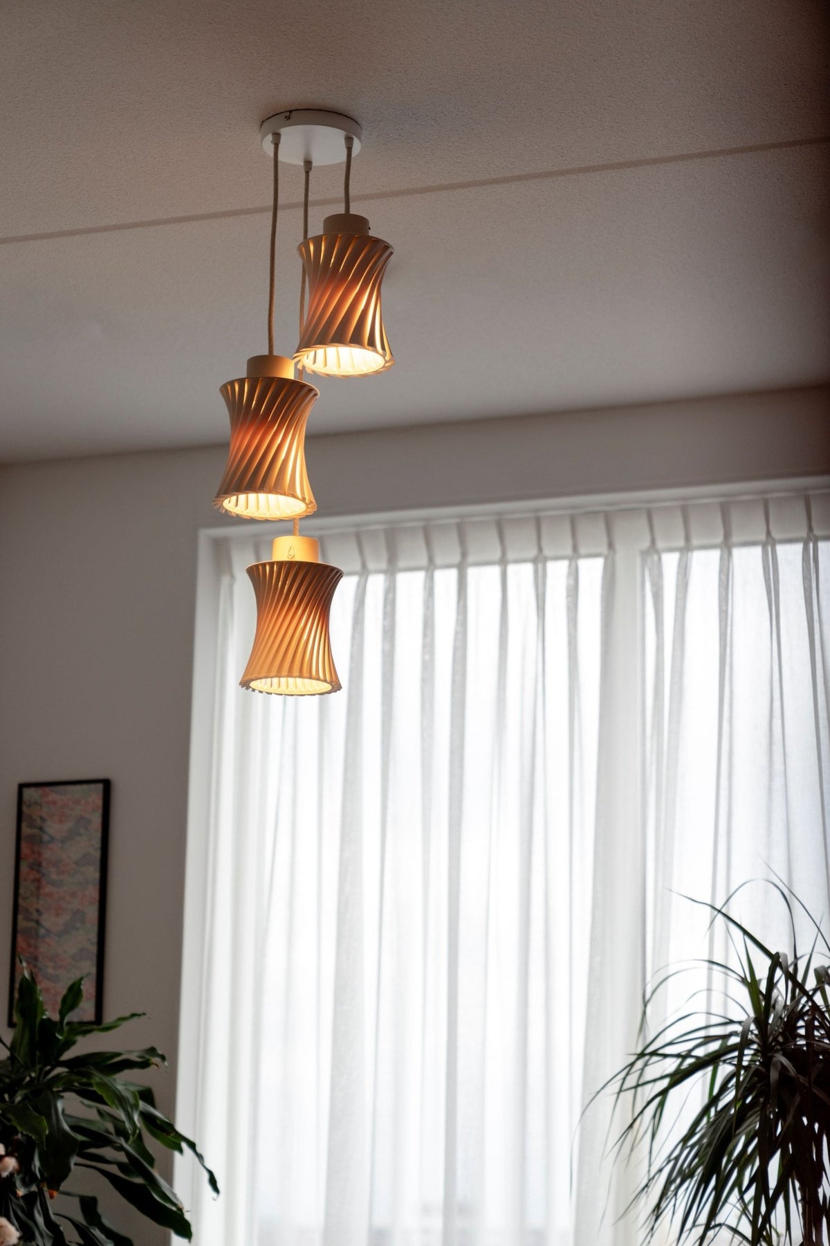 Drie hanglampen getrapt onder een plafond in een woonkamer met planten en een raam met witte gordijnen.