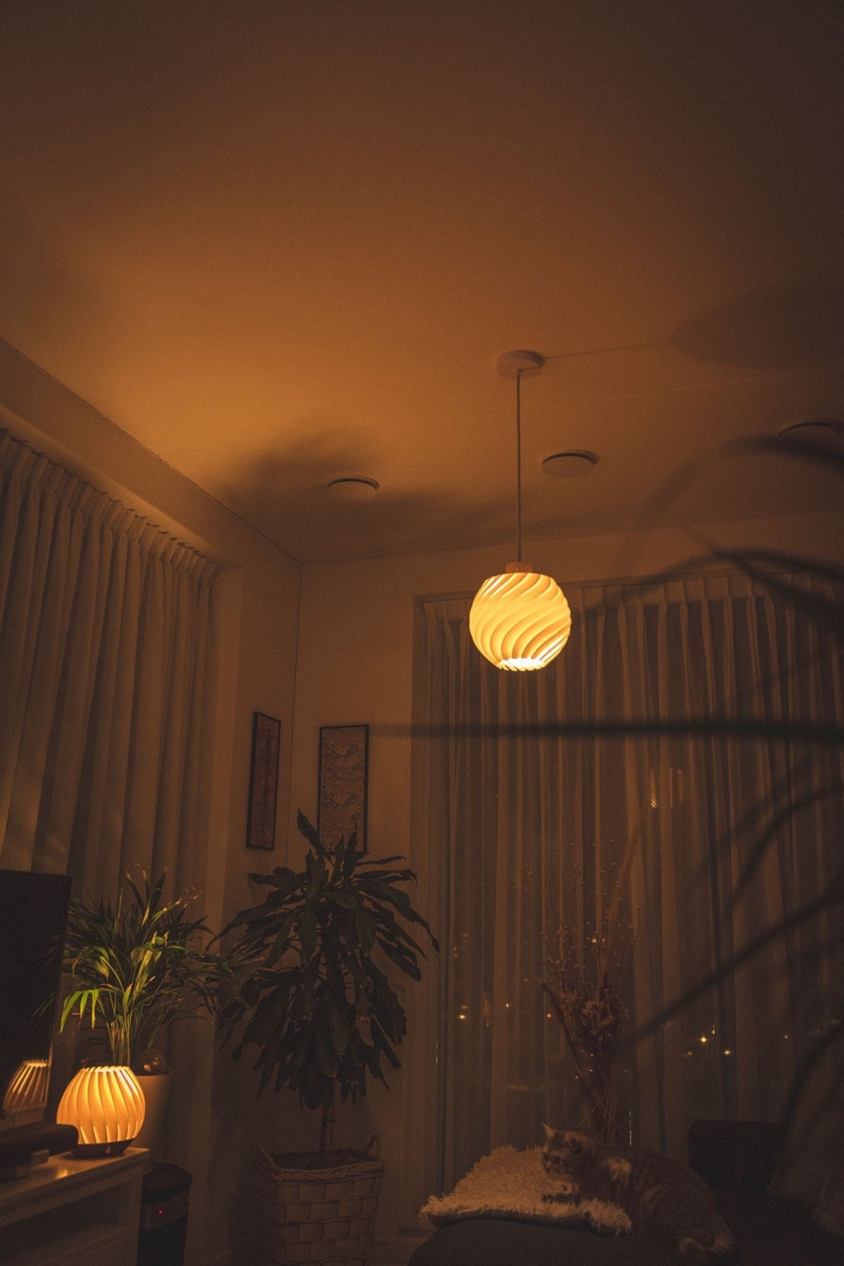 Brandende bolvormige hanglamp in een schemerige woonkamer met planten en gordijnen.