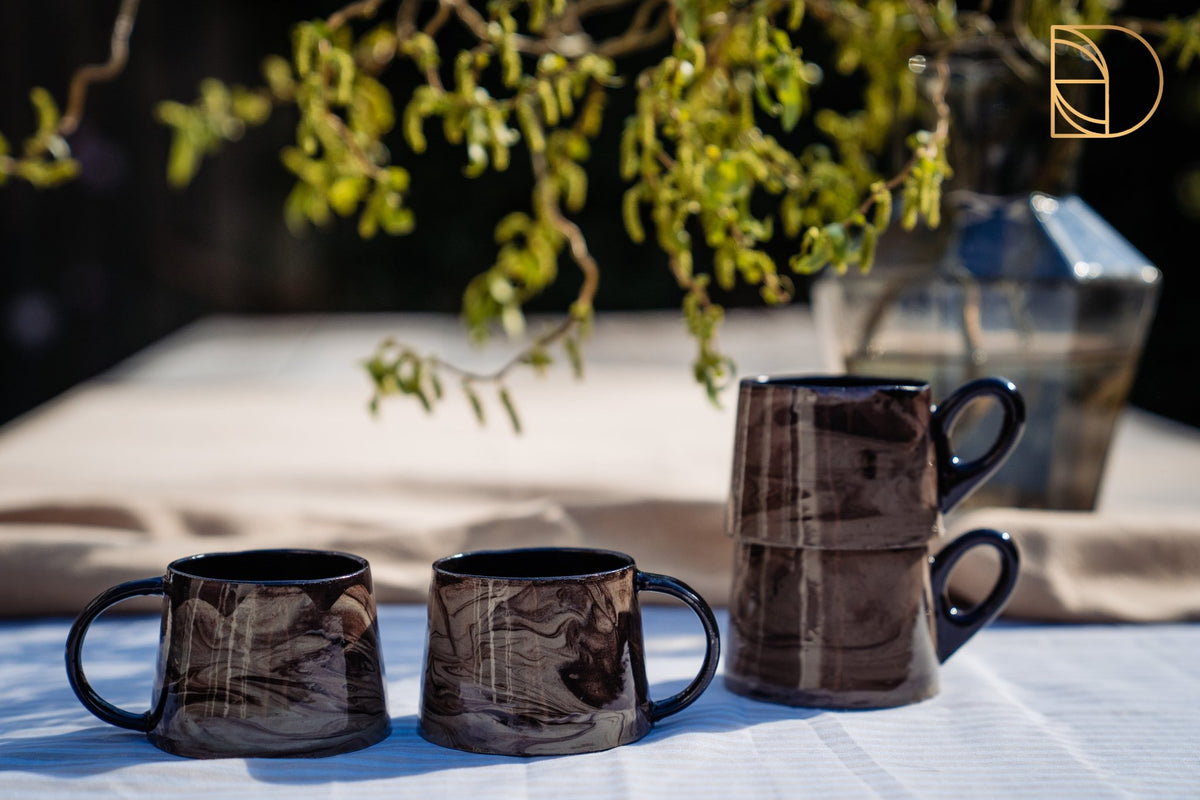 Drie keramieken mokken met bruin-zwart gemarmerd glazuur op een tafel in de tuin.