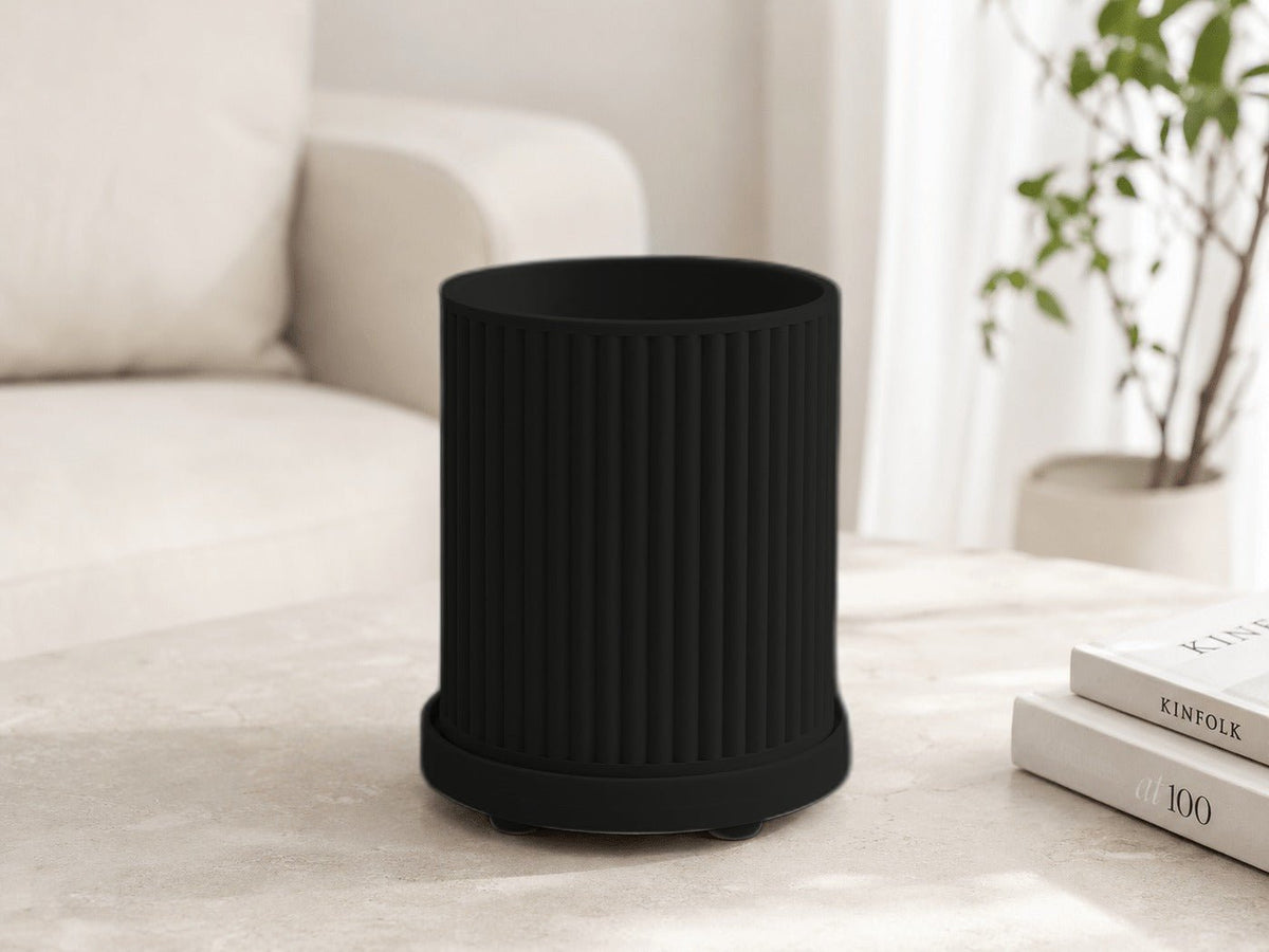 Geribbelde plantenpot in mat zwart met onderschotel, op een tafel bij een bank.