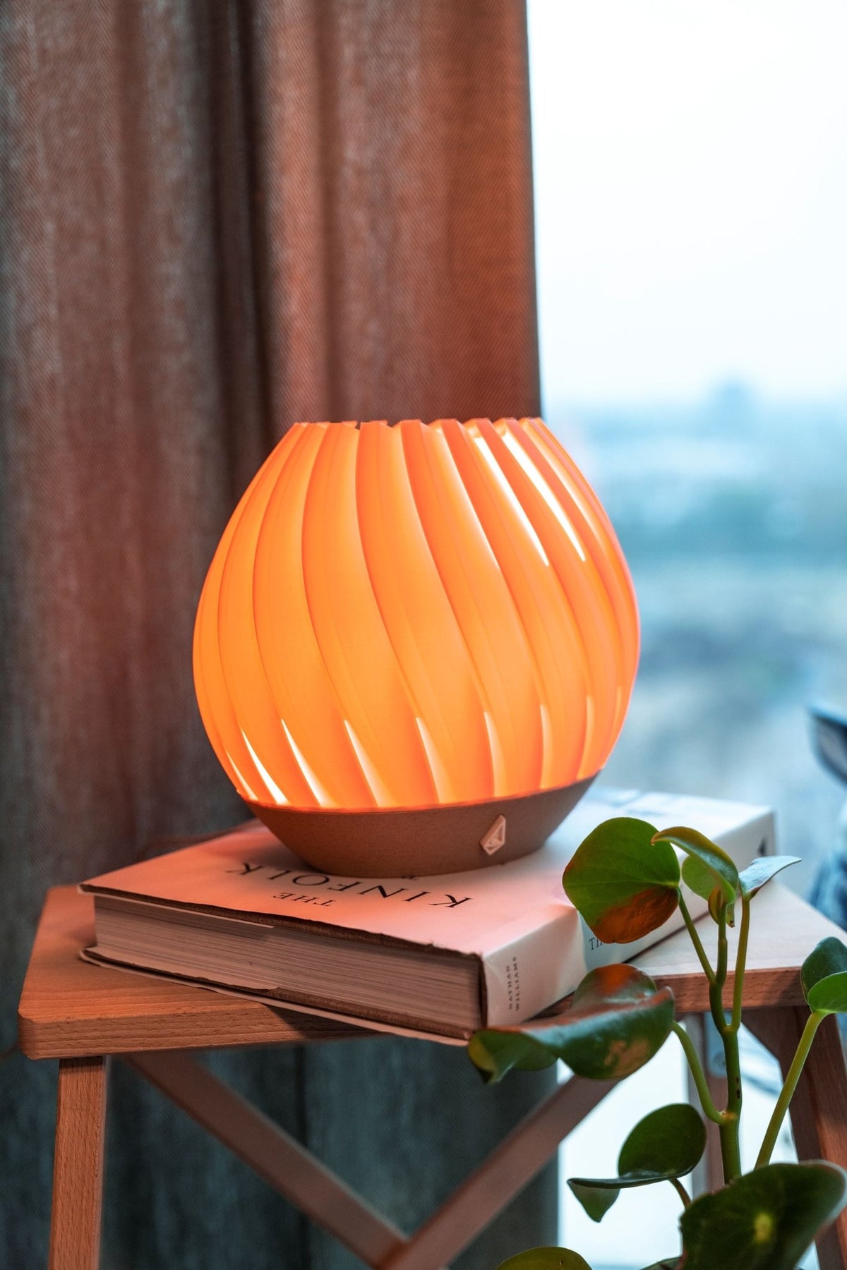 Koraalroze brede tafellamp met brandend licht op boeken op een houten bijzettafel bij een raam.