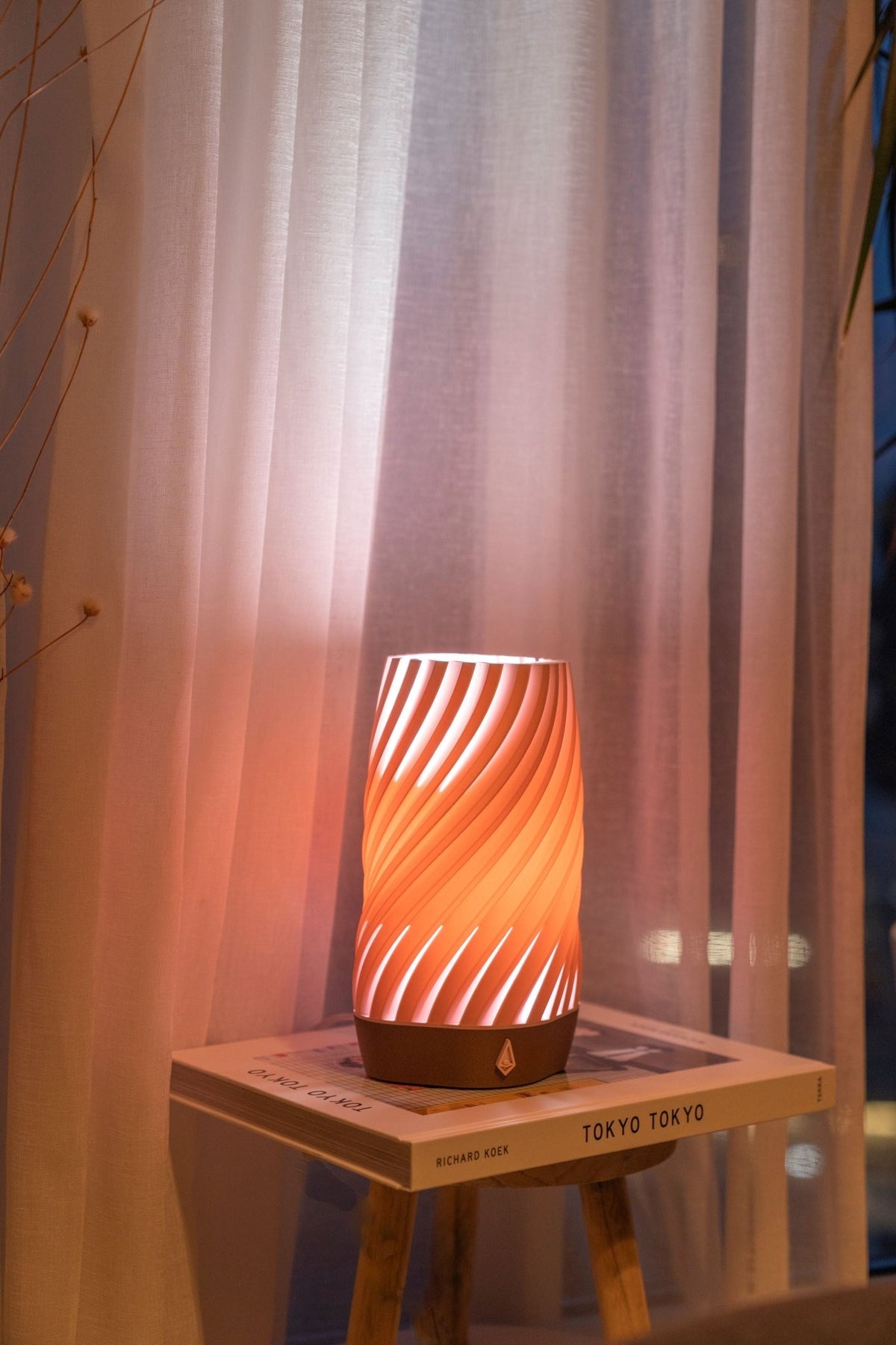 Koraalroze smalle tafellamp brandt op een houten bijzettafel voor een gordijn in avondlicht.