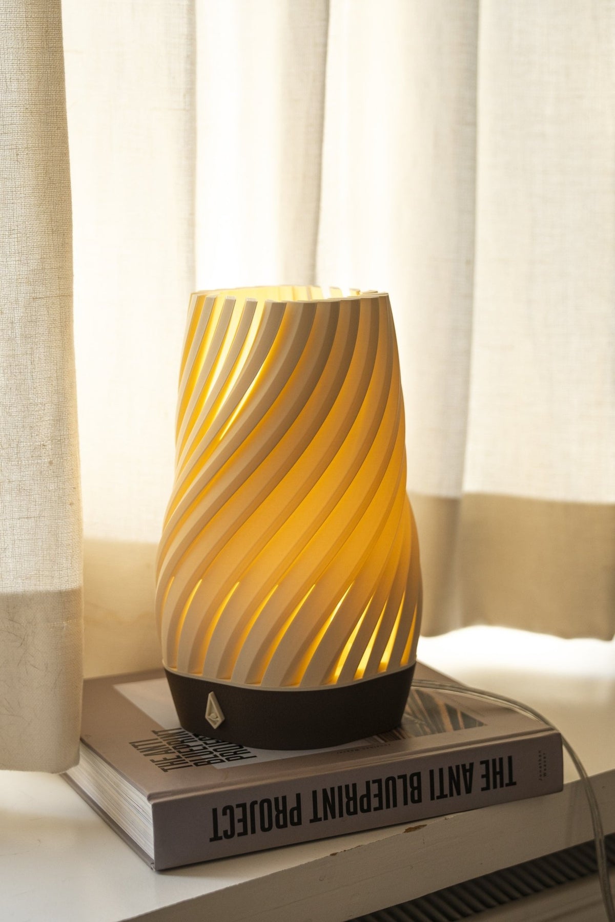 Smalle gedraaide tafellamp met warm brandend licht op een stapel boeken voor een licht gordijn.
