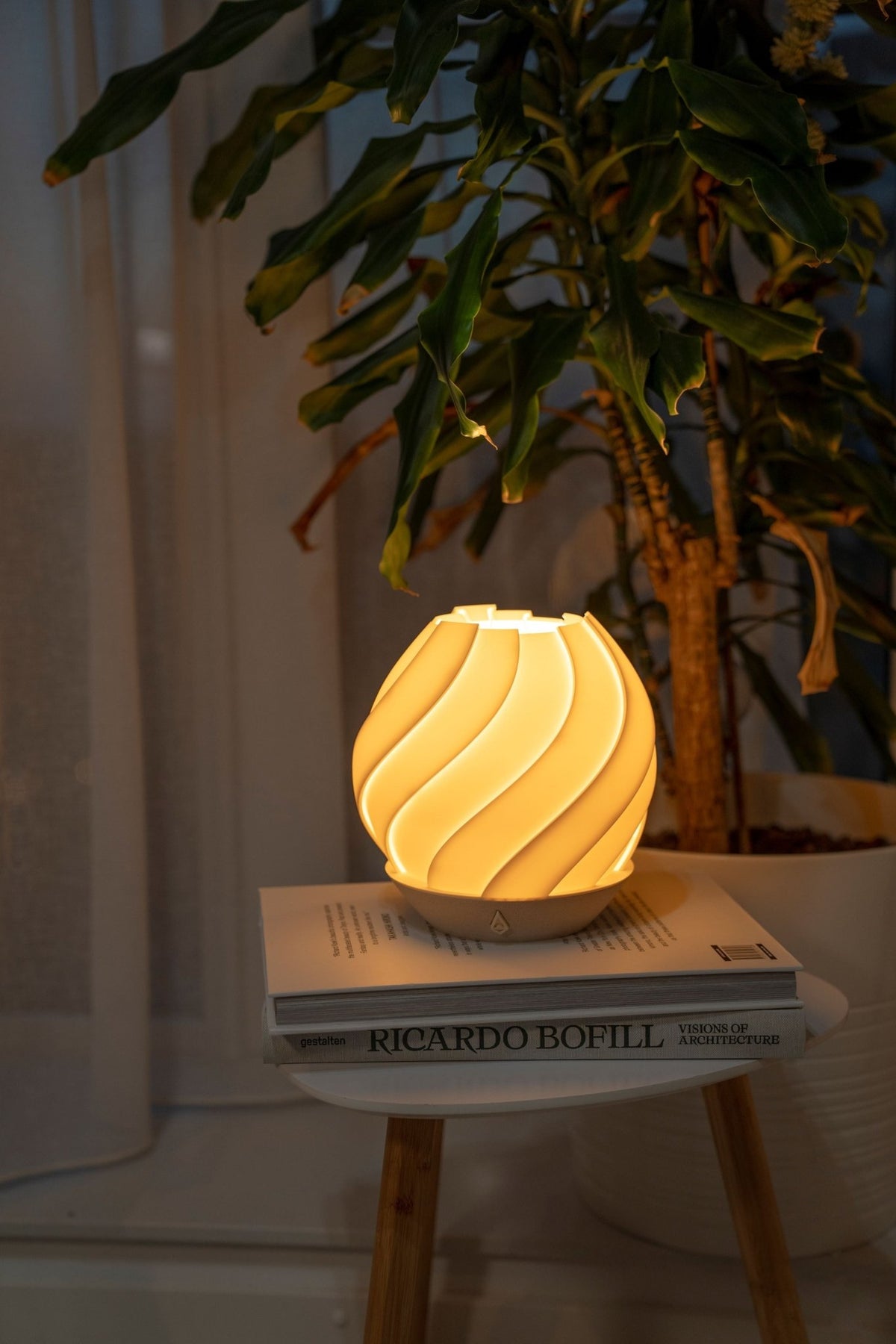 Brandende bolvormige tafellamp op een boek naast een kamerplant voor een licht gordijn.