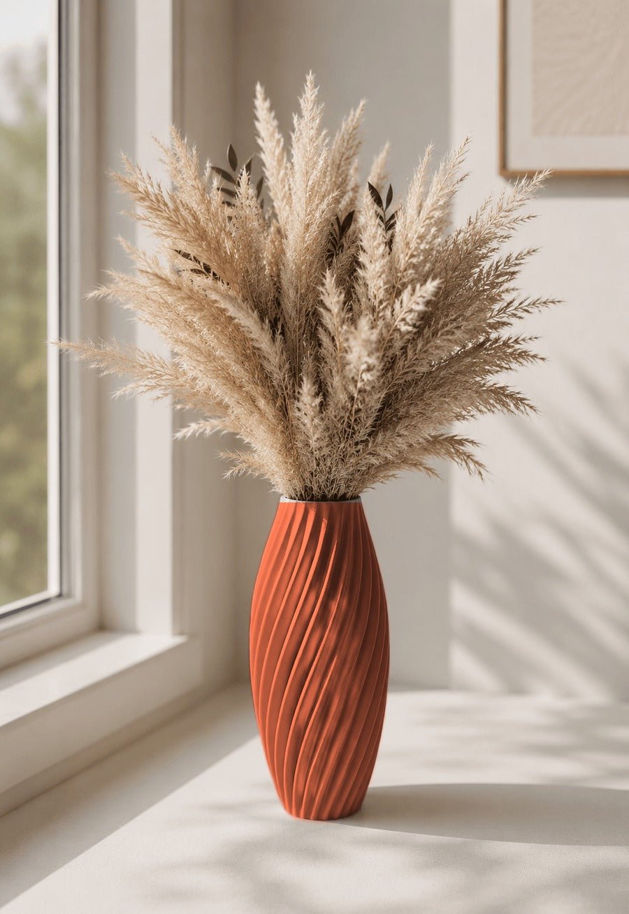 Slanke gedraaide vaas in terracotta met pampaspluimen erin, bij een raam.