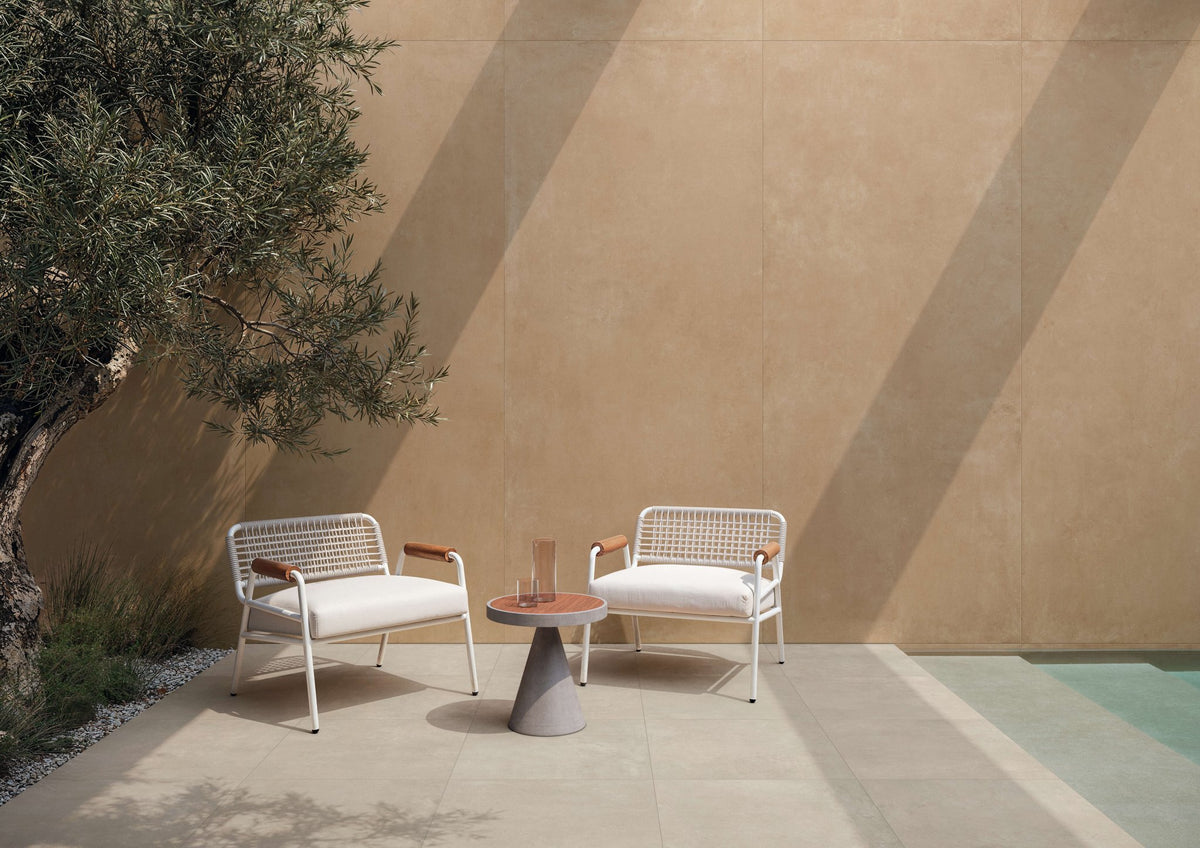 Terras met zandkleurige betonlook tegels, twee gevlochten fauteuils en een olijfboom.