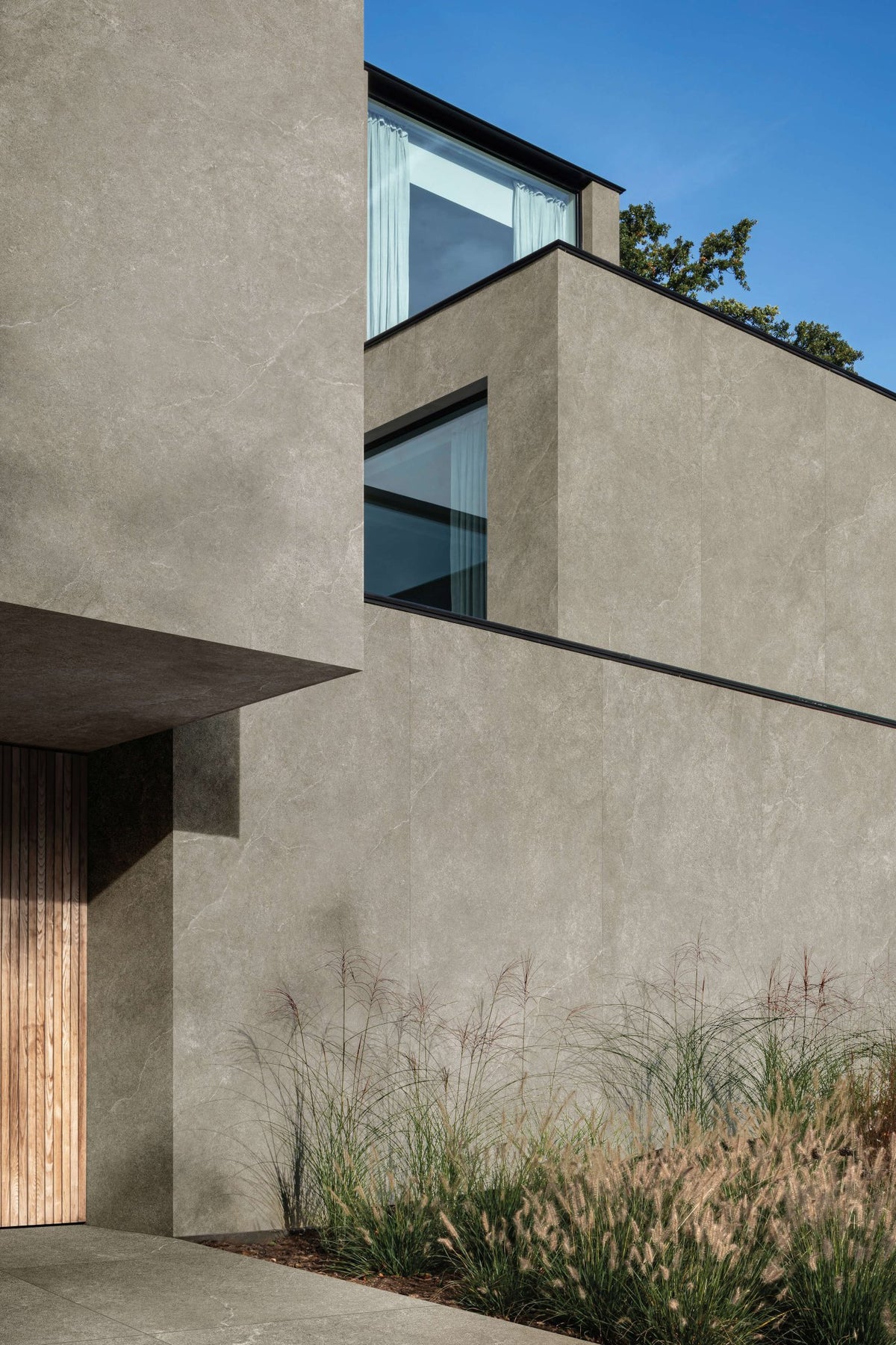 Moderne gevel met taupe kalksteenlook tegels, houten voordeur en siergrassen.